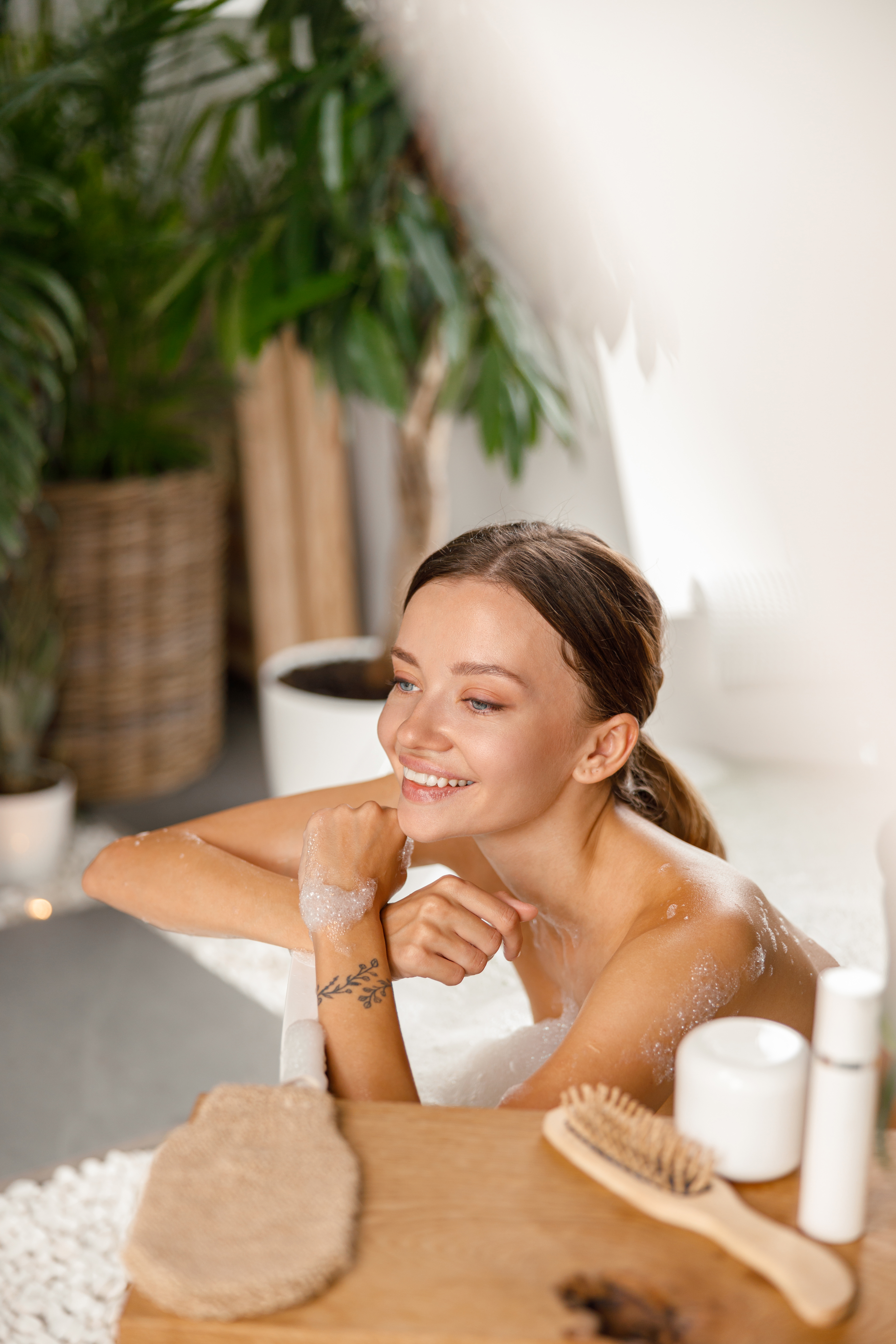 Joyful young woman smiling away and leaning on bathtub side while bathing at spa resort